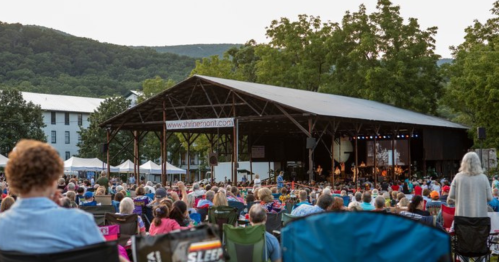 A large crowd enjoys a concert at an outdoor stage surrounded by trees and mountains.