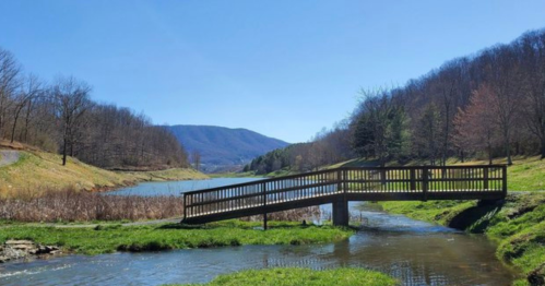 A wooden bridge spans a stream, surrounded by green grass and trees, with mountains in the background under a clear blue sky.