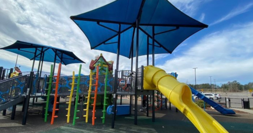 Colorful playground with slides and climbing structures, shaded by large blue canopies under a cloudy sky.