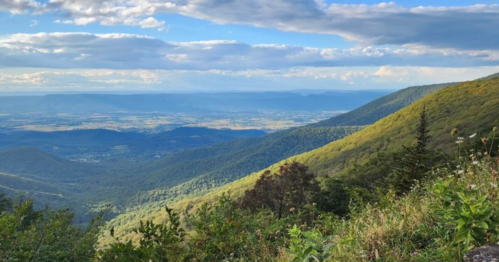 A panoramic view of lush green mountains and valleys under a blue sky with scattered clouds.