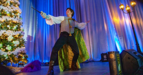 A performer in a cape poses dramatically with a wand on stage, with a decorated Christmas tree in the background.