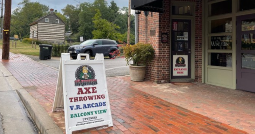 A sidewalk sign advertising axe throwing, VR arcade, and balcony view at a business entrance on a rainy day.