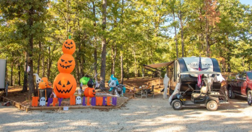 A festive Halloween display with stacked pumpkin decorations and ghost figures in a wooded campsite.