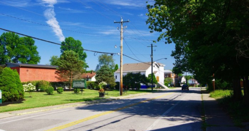 A sunny street scene with green trees, power lines, and buildings on either side, leading into a clear blue sky.