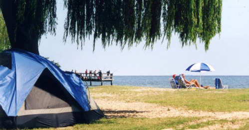 A blue tent on a sandy beach with a fishing pier and a person relaxing under an umbrella by the water.