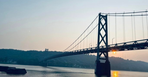 A suspension bridge spans a river at sunset, with hills in the background and a calm water surface reflecting the light.