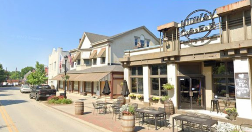 A quaint street scene featuring a coffee shop and shops with outdoor seating and planters under a clear blue sky.
