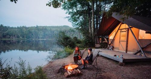 Two people sit by a campfire near a tent by a calm lake, surrounded by trees at dusk.