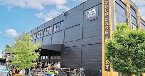 A modern brewery with outdoor seating, surrounded by trees and bicycles, under a blue sky.