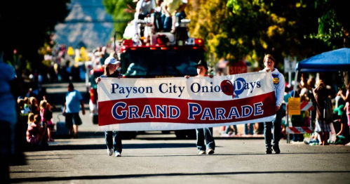 Two people hold a large banner reading "Payson City Onion Days Grand Parade" in a festive street scene.