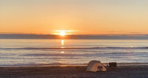 A serene beach at sunset, with a tent and a chair on the sand, reflecting golden hues on the calm ocean.