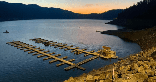 A serene lake at sunset with a boat dock and rocky shoreline, surrounded by mountains and trees.