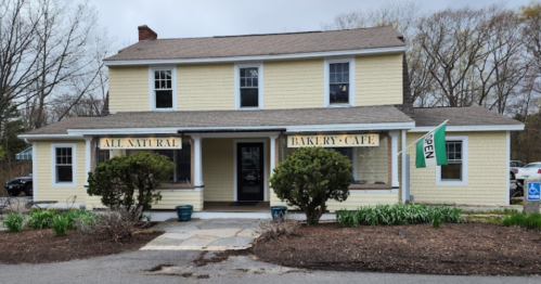 A yellow house with a sign reading "All Natural Bakery-Cafe" and an "Open" flag outside, surrounded by greenery.