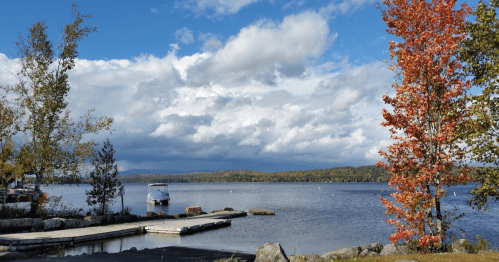 A serene lake scene with a wooden dock, colorful autumn trees, and a cloudy blue sky in the background.