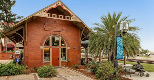 Historic brick train station with palm trees and a statue nearby, set against a clear blue sky.
