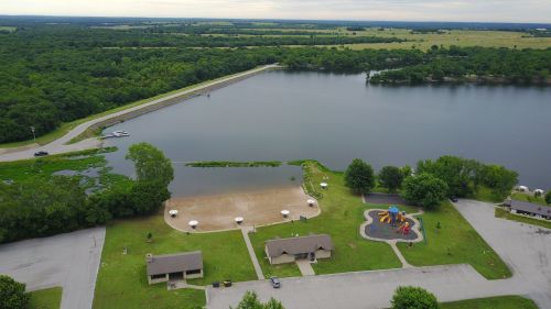 Aerial view of a lake with a playground, picnic area, and green landscape under a cloudy sky.