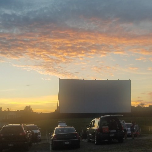 A drive-in movie screen silhouetted against a colorful sunset, with parked cars in the foreground.