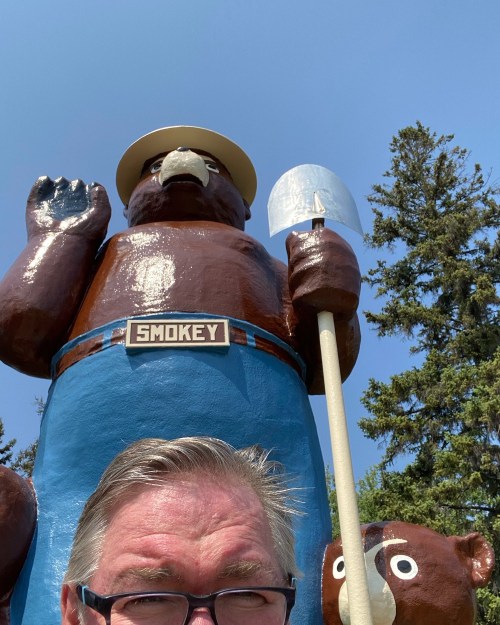 A person takes a selfie in front of a large Smokey Bear statue holding a shovel, surrounded by trees.