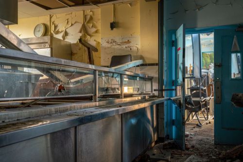 An abandoned restaurant interior with broken glass, scattered debris, and peeling walls, illuminated by natural light.
