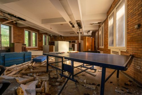 An abandoned room with a blue table, scattered debris, and sunlight streaming through large windows.