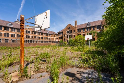 Abandoned basketball court overgrown with weeds, surrounded by old brick buildings under a clear blue sky.
