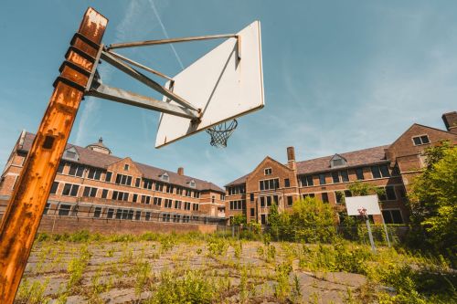 Abandoned basketball court with overgrown weeds and old brick buildings in the background under a clear blue sky.