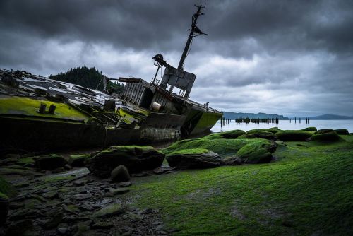 A shipwreck covered in green moss rests on a rocky shore under a cloudy sky, with calm waters in the background.
