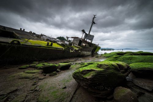 An abandoned ship rests on rocky shore, covered in moss, under a cloudy sky.