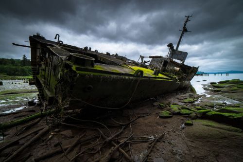 A weathered shipwreck covered in moss rests on a rocky shore under a cloudy sky.