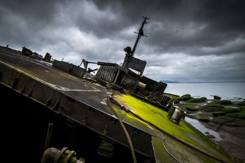 A shipwreck tilted on rocky shore, covered in green moss, under a dramatic, cloudy sky.