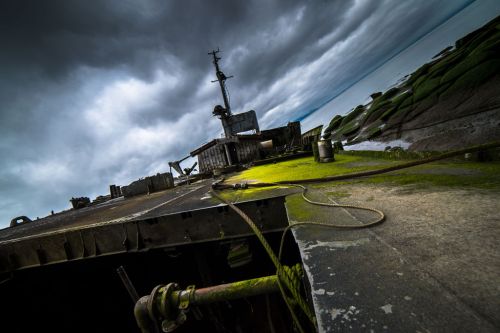 A weathered shipwreck covered in green moss, set against a dramatic cloudy sky and calm waters.