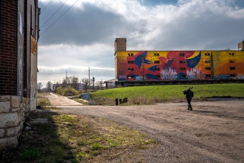 A person walks along a gravel path beside a colorful graffiti-covered building under a cloudy sky.