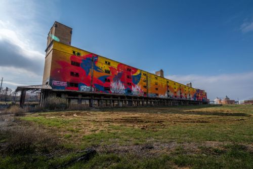 A large, colorful mural covers the side of an abandoned industrial building against a clear blue sky.