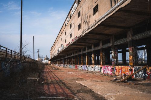 Abandoned industrial building with graffiti, overgrown vegetation, and a clear blue sky in the background.