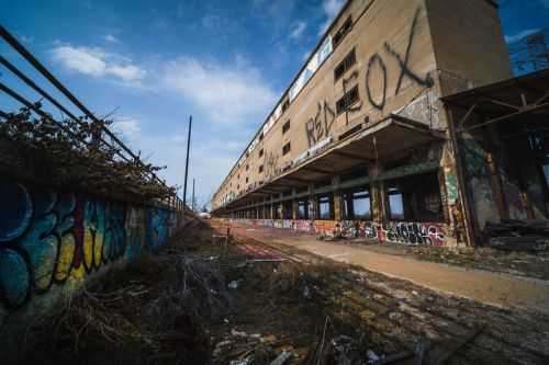 Abandoned building with graffiti, overgrown vegetation, and a cloudy sky in the background.