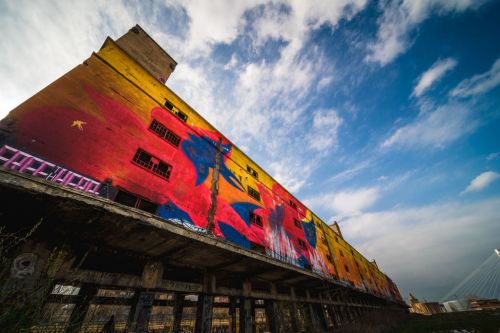 A colorful mural on a tall, abandoned building under a blue sky with scattered clouds.