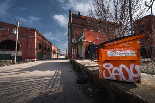 An orange dumpster sits on a sidewalk in an urban alley, surrounded by red brick buildings and graffiti under a blue sky.