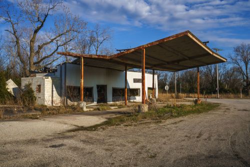 Abandoned gas station with a rusted canopy, overgrown vegetation, and a gravel road under a blue sky.
