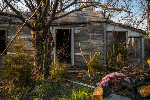 Abandoned house with peeling paint, overgrown vegetation, and debris scattered in front, illuminated by warm sunlight.