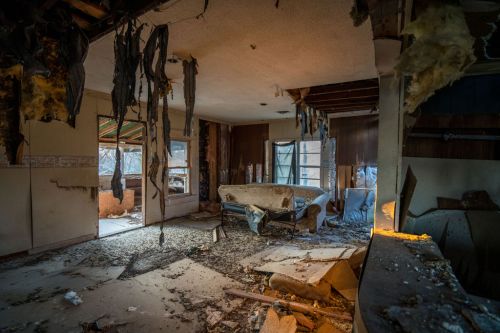 A dilapidated room with a damaged ceiling, debris on the floor, and a worn-out couch amidst signs of neglect and decay.