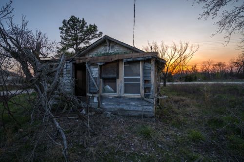 Abandoned wooden house with broken windows, surrounded by overgrown grass and trees at sunset.