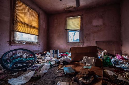 A cluttered, abandoned room filled with trash, old furniture, and sunlight streaming through dusty windows.