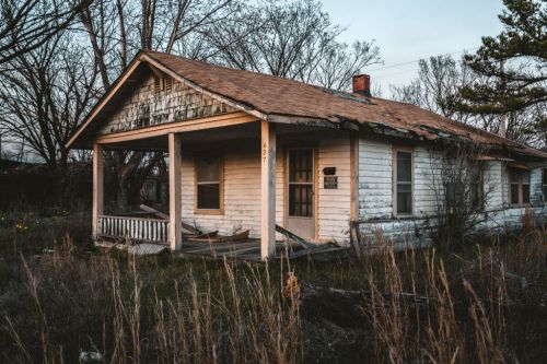 Abandoned white house with a sagging roof, overgrown grass, and trees in a desolate setting during twilight.