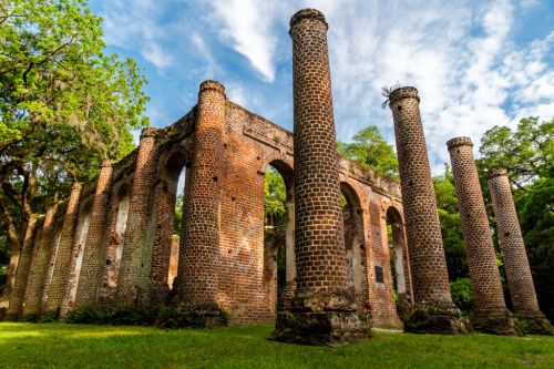 Ruins of a brick structure with tall columns, surrounded by greenery and under a blue sky with scattered clouds.