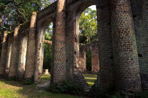 Ruins of a brick structure with arched openings, surrounded by greenery and trees in a serene outdoor setting.