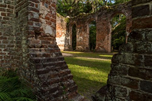 Ruins of a brick structure surrounded by greenery, with arches and moss-covered walls in a serene outdoor setting.