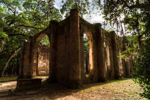 Ruins of a brick structure surrounded by lush greenery and trees, with sunlight filtering through the foliage.