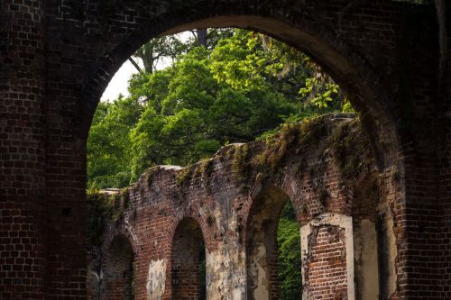 Ruins of a brick structure with arched openings, surrounded by lush green trees and foliage.