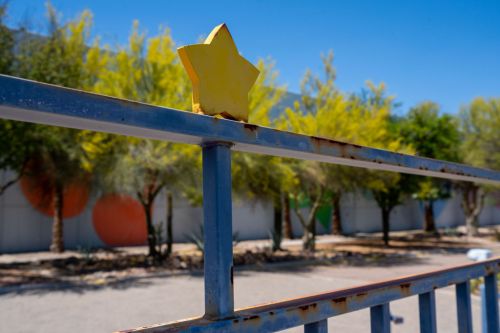 A close-up of a yellow star decoration on a rusty metal railing, with trees and colorful circles in the background.