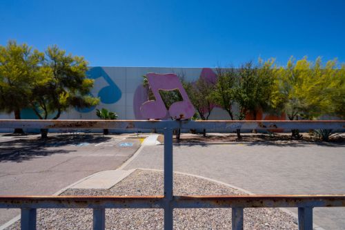 A pink musical note sculpture on a rusted fence in front of a colorful mural and desert vegetation under a clear blue sky.